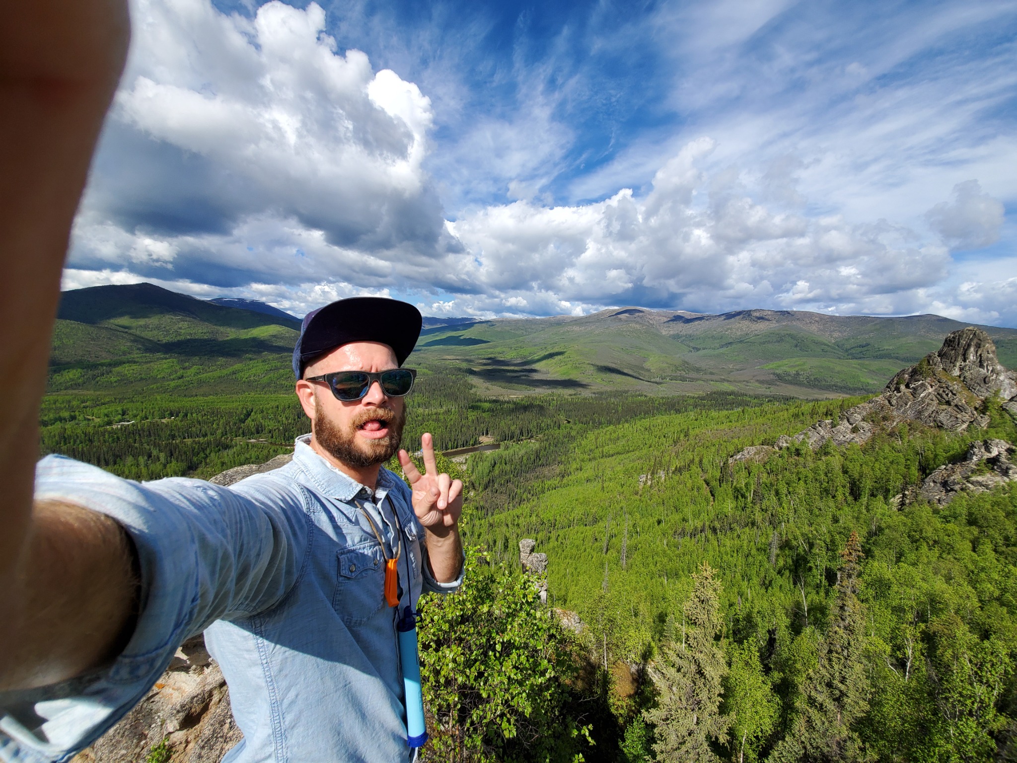 Yorudan taking a selfie above a green mountain valley beneath a wide, cloud-filled sky.